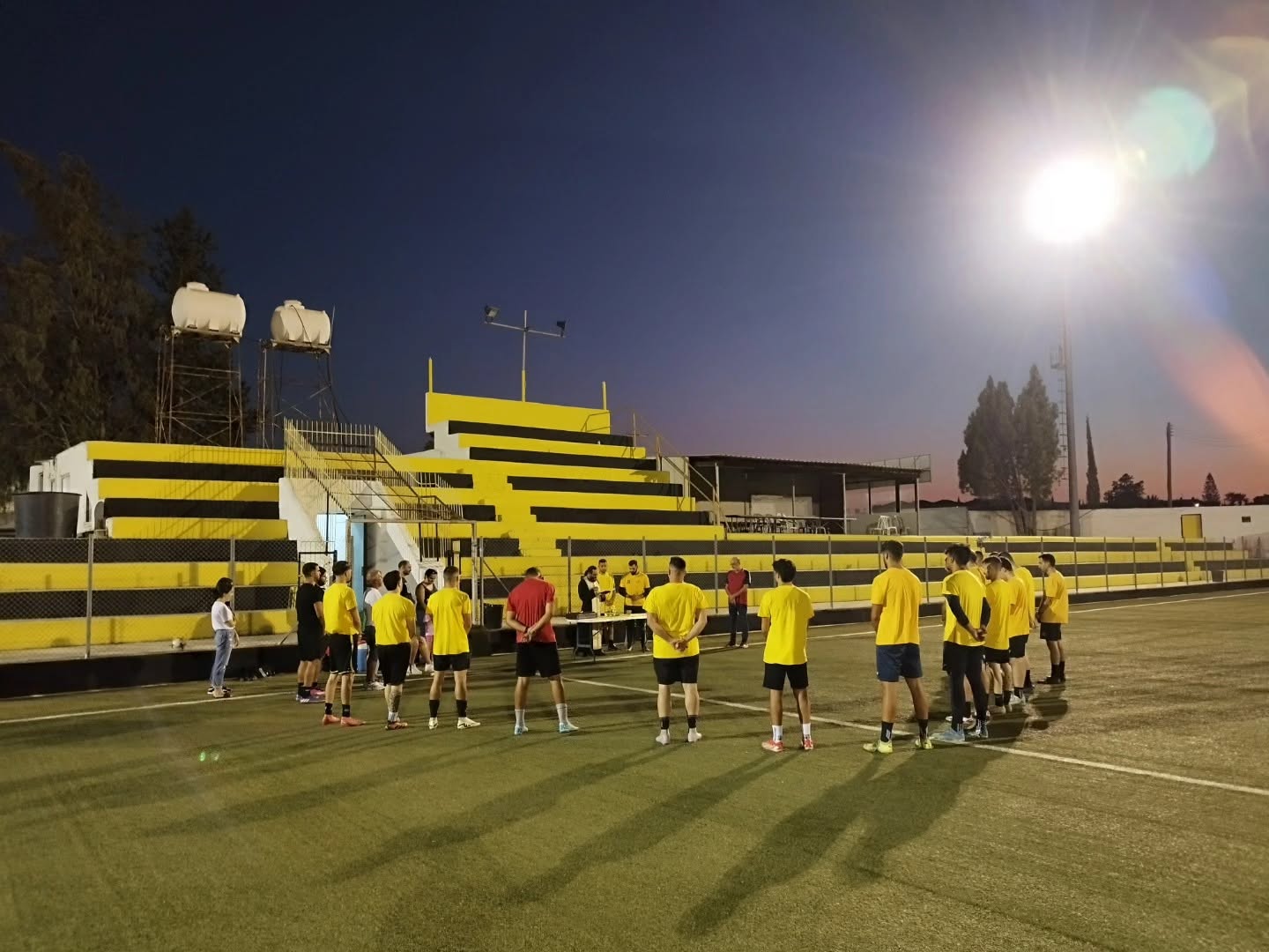 Players in yellow shirts standing on the pitch in front of the yellow and black stands at dusk