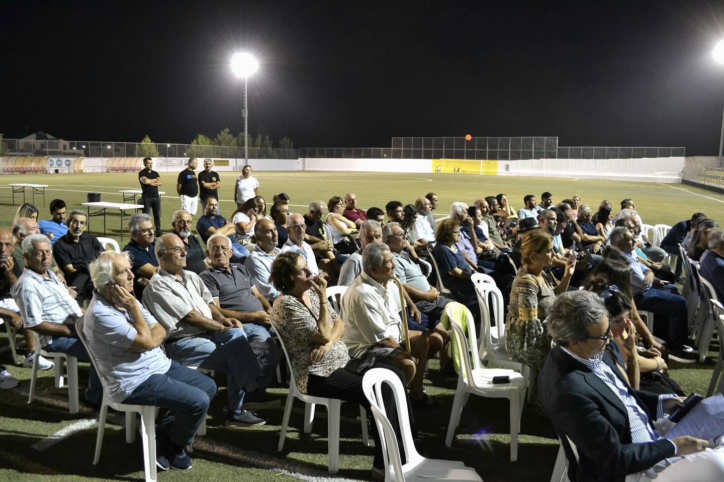 Members of the community seated on the pitch under floodlights during an evening gathering
