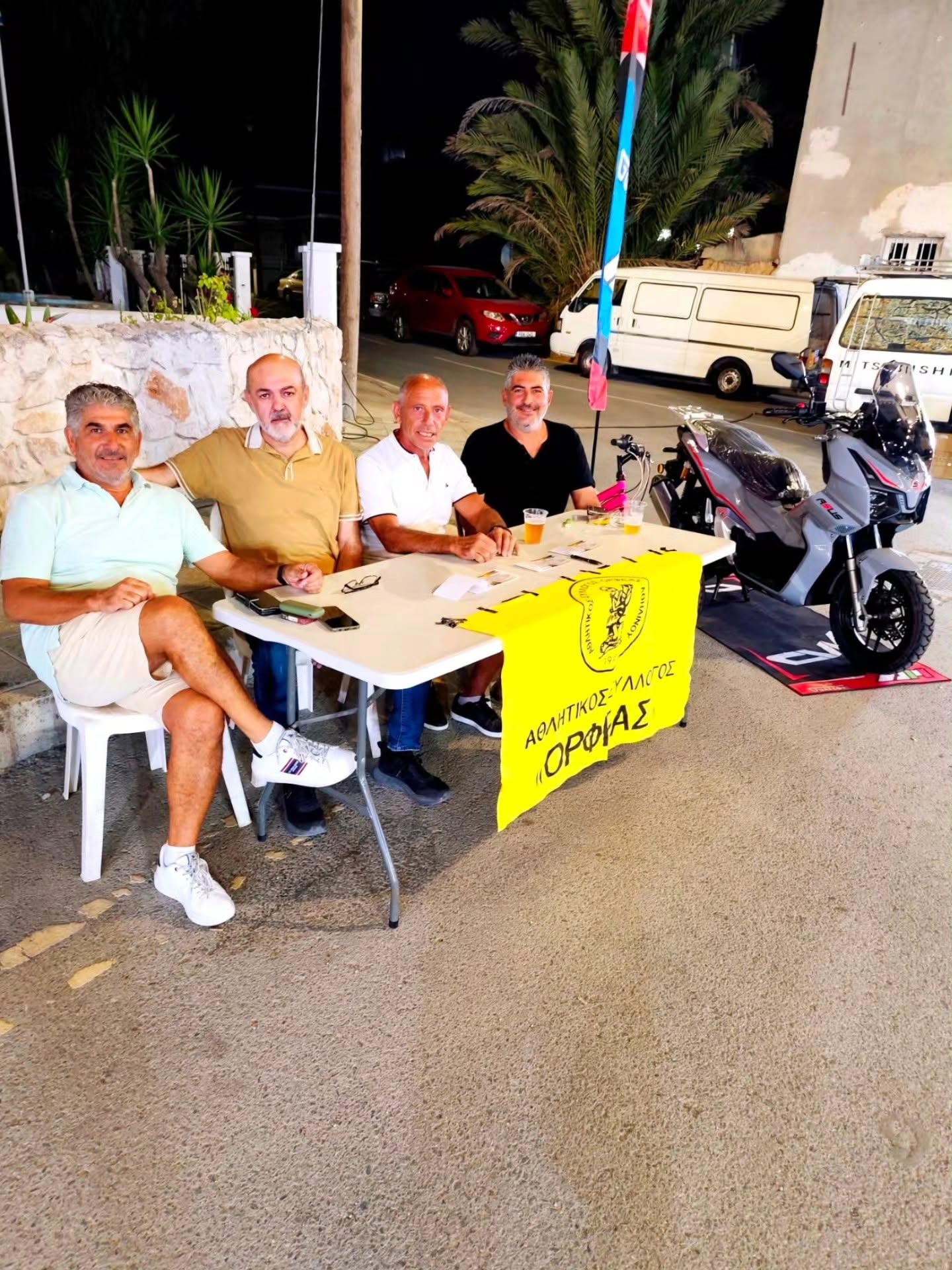 Four members seated at a table with the association's yellow banner, outdoors in the evening